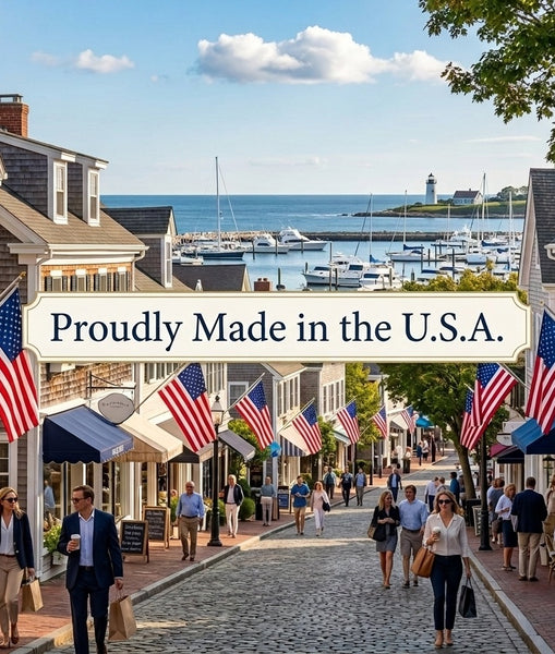 A scenic coastal town street lined with shops and American flags, with people walking toward the harbor and boats on the water. A banner across the center reads 'Proudly Made in the U.S.A.